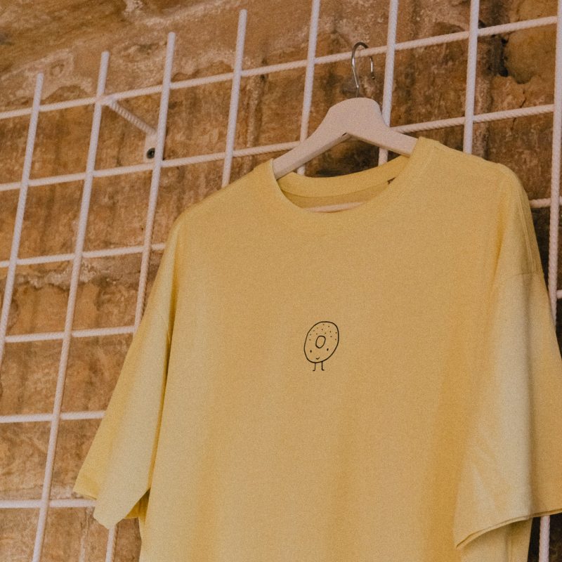 "100% cotton boxy t-shirt featuring a casual fit, displayed on a hanger against a stone background showcasing the bagel shop's logo."